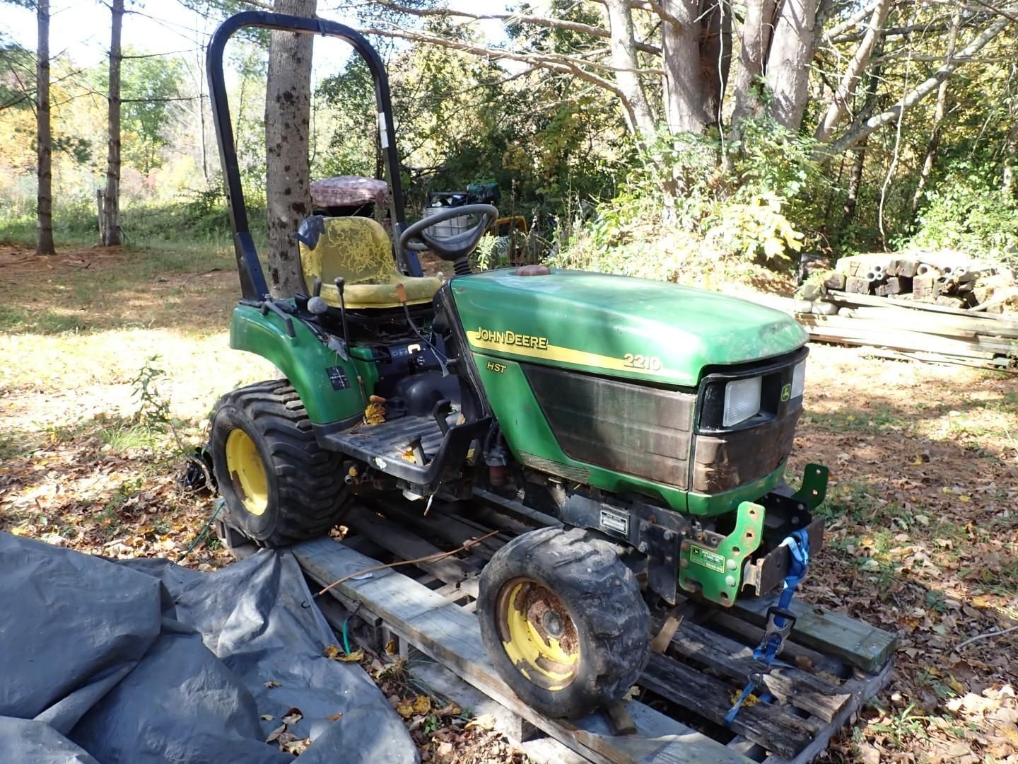 Green tractor parked on a black tarp in a wooded yard, with a roll bar and front loader attachments.