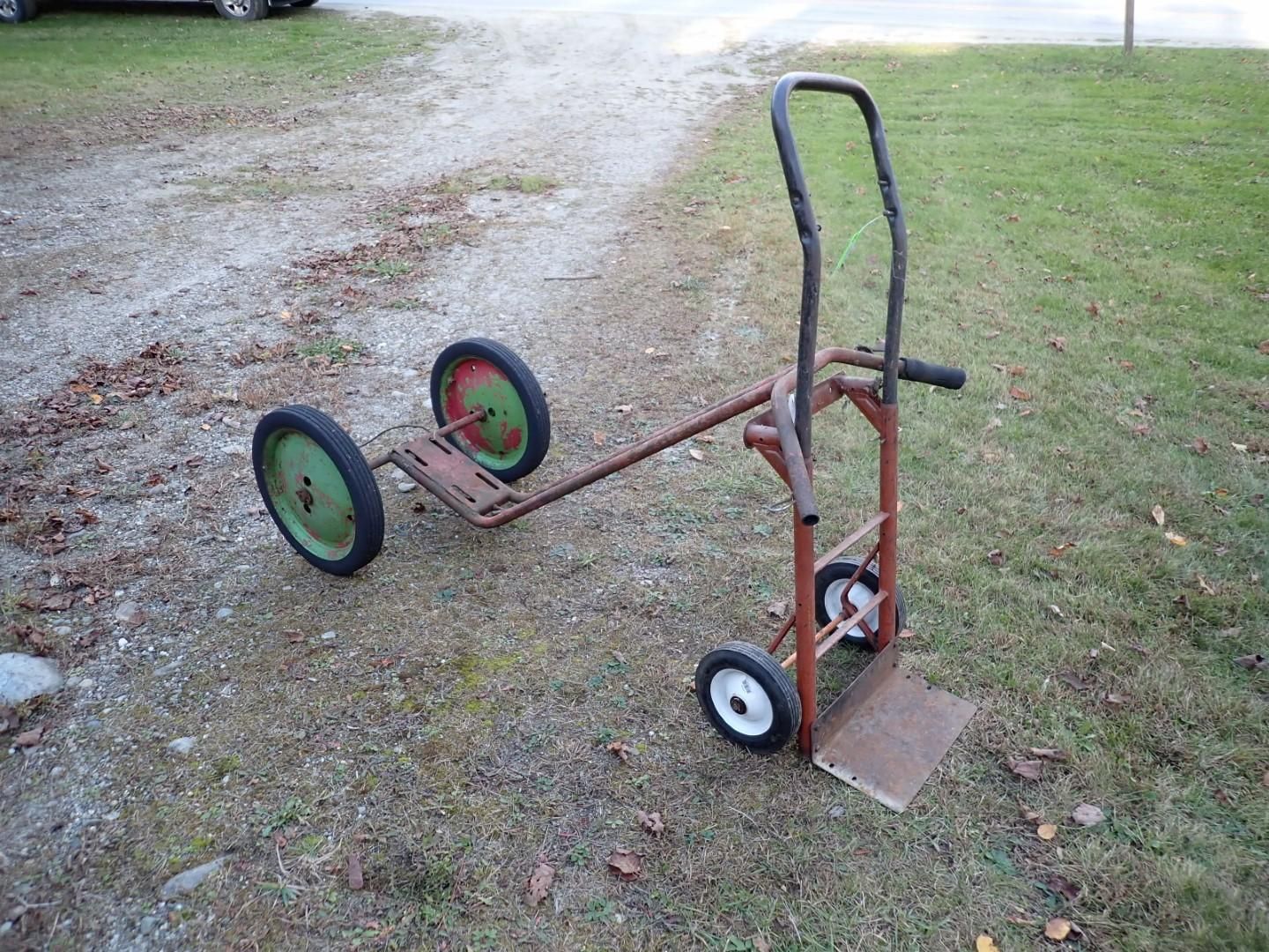 Metal hand truck with two large green wheels and two small white wheels on a grassy area.