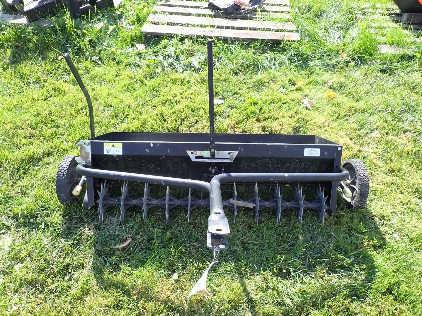 Black garden tiller attachment on grass, with metal tines and wheels under a wooden handle.