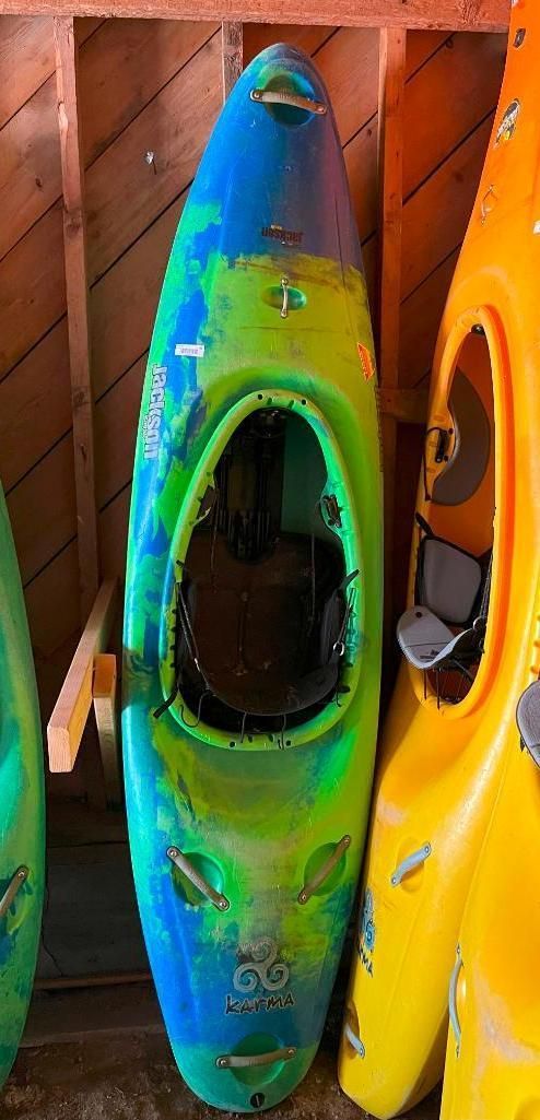 A colorful kayak in blue, green, and yellow leans against a wooden wall. Another yellow kayak is next to it.