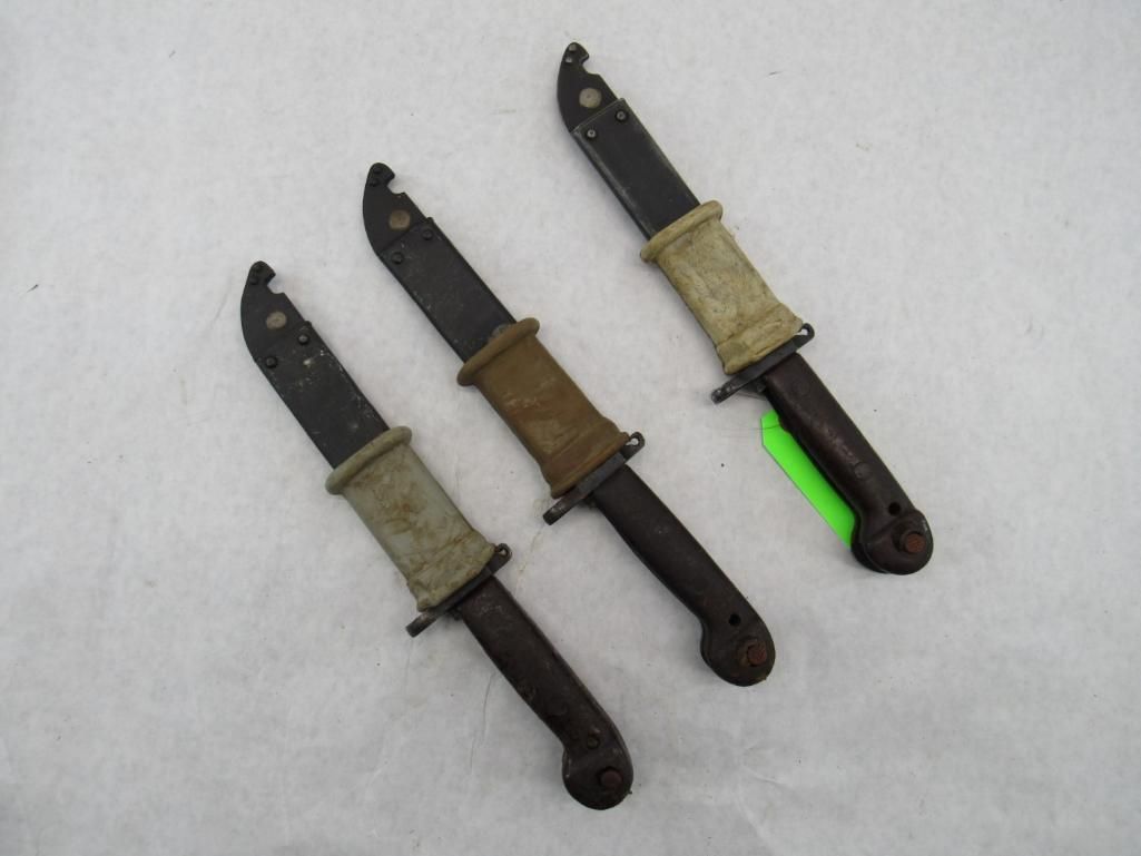 Three vintage military bayonets on a white surface, blades facing up, with tan and brown handles.