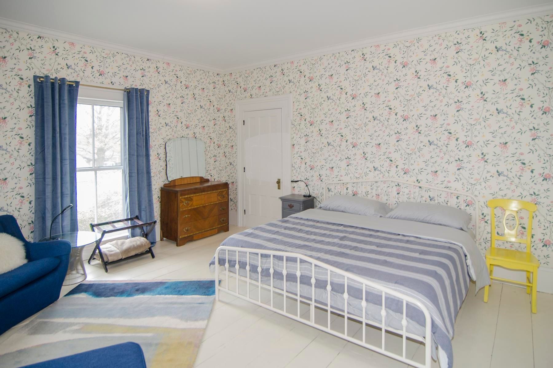 Bedroom with patterned wallpaper, white bed frame, blue curtains and armchair, and yellow chair.