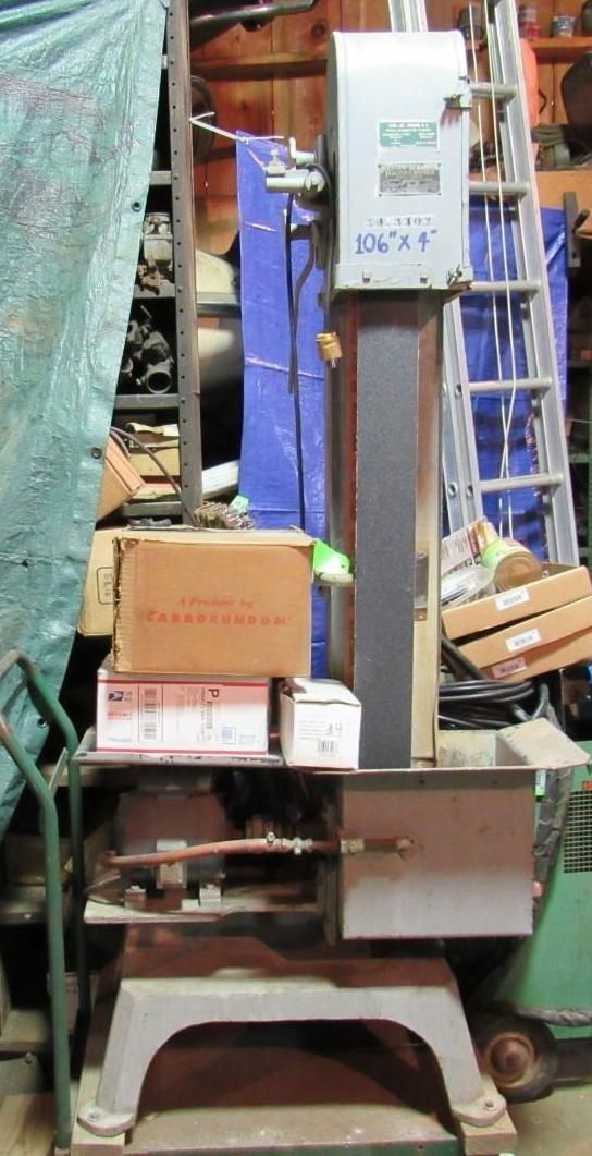 Belt sander machine in a workshop setting. Silver, beige, and gray metal. Boxes stacked on a lower shelf.