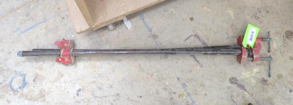 A long bar clamp lying on a concrete surface, with a red clamp at each end.