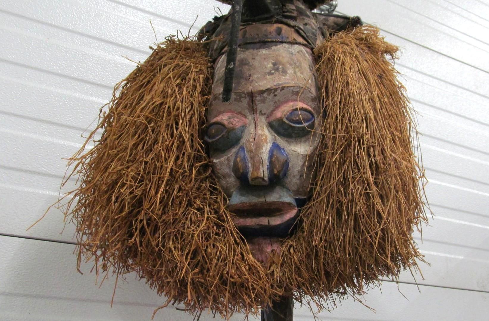Wooden African mask with straw-like fringe, painted face with blue and red details, against a white wall.