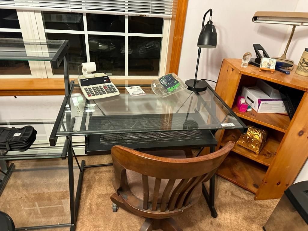 Glass-top desk with keyboard and calculator, wooden chair, lamp, and bookshelf near a window.