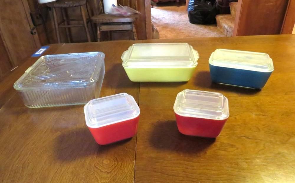 Five vintage food storage containers on a wooden table; clear glass, yellow, blue, and two red containers with white lids.
