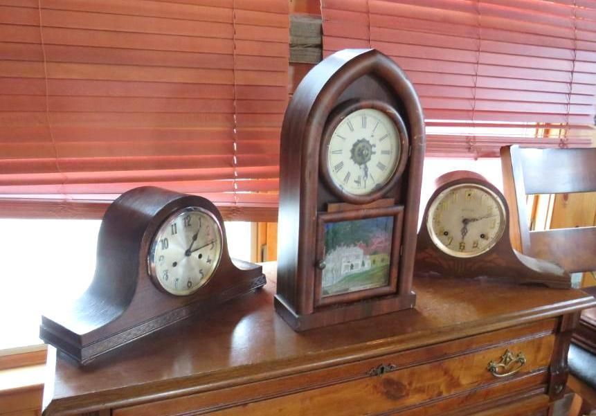 Three antique wooden clocks on a wooden dresser, in front of a window with blinds.