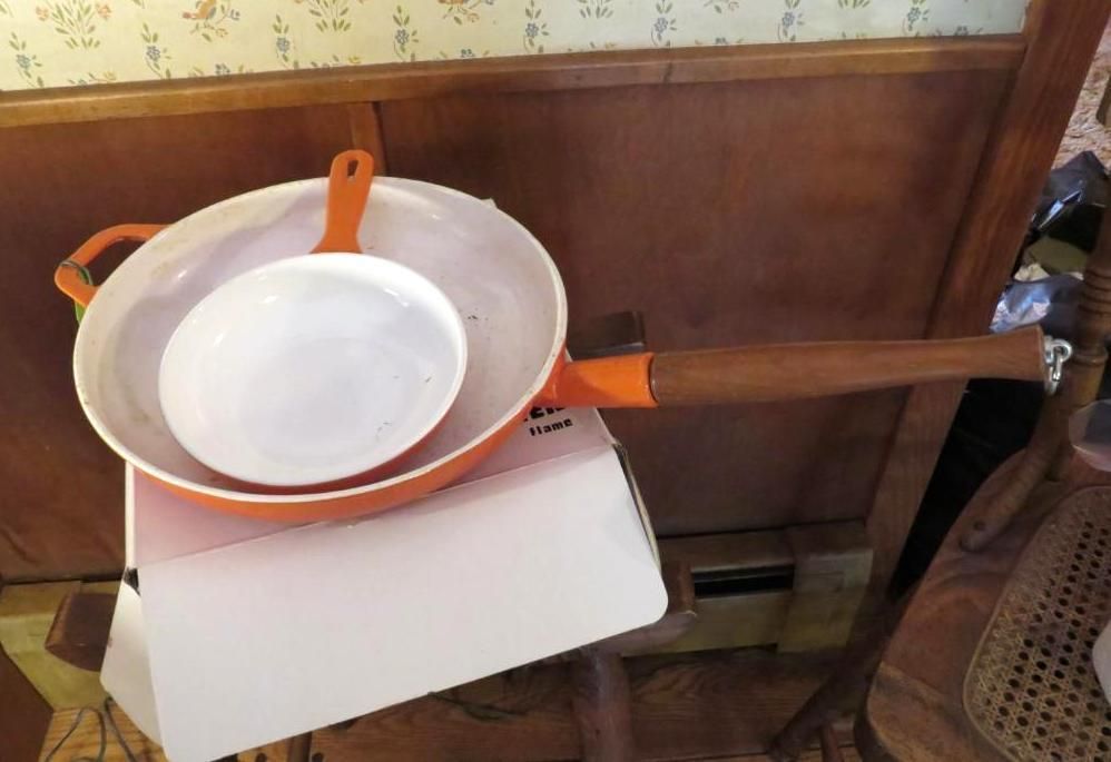 Orange and white enameled pans stacked on a box, wood handle, indoor setting.