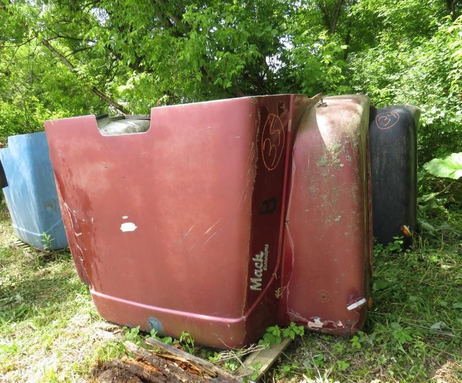 Burgundy Mack truck hood, outdoors, resting in grass. Other colored truck parts nearby.