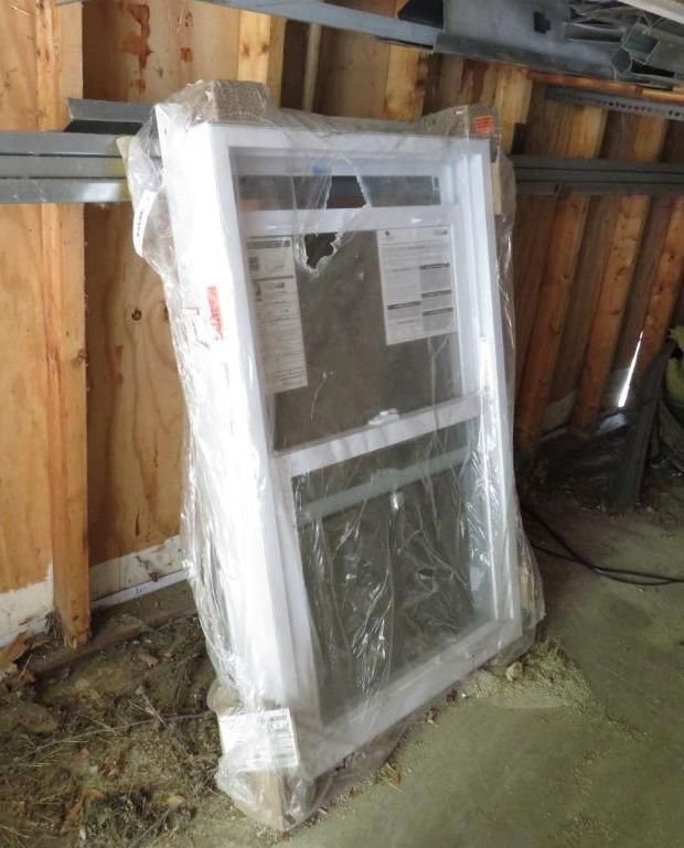 New, white window in protective plastic wrap, leaning against a wooden wall in a construction setting.