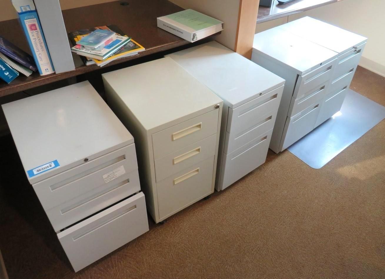 Four metal filing cabinets of varying sizes in a room, under a desk with some books and office supplies on top.