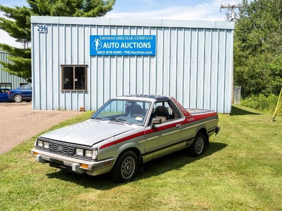 Silver Subaru Brat pickup truck parked in front of a blue-gray building with an 