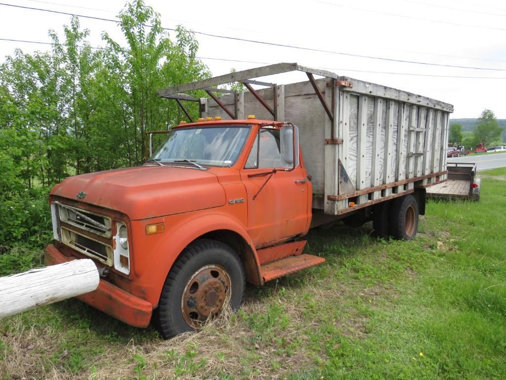 Orange vintage truck with a large, light-colored bed parked on grassy roadside.