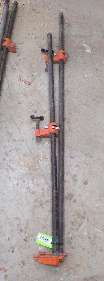 Two pipe clamps on a concrete surface. The clamps have black pipes and orange clamp parts. A green tag is attached.