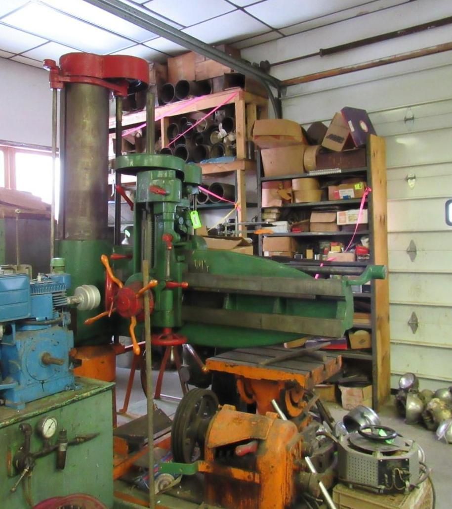 A large, green industrial drill press in a workshop with shelves of supplies.