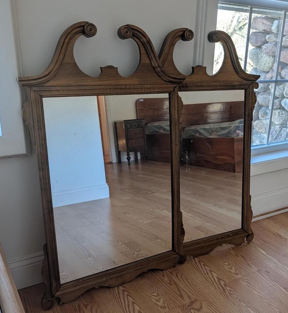 Ornate wooden double mirror with curved top, standing on a hardwood floor by a bright window.