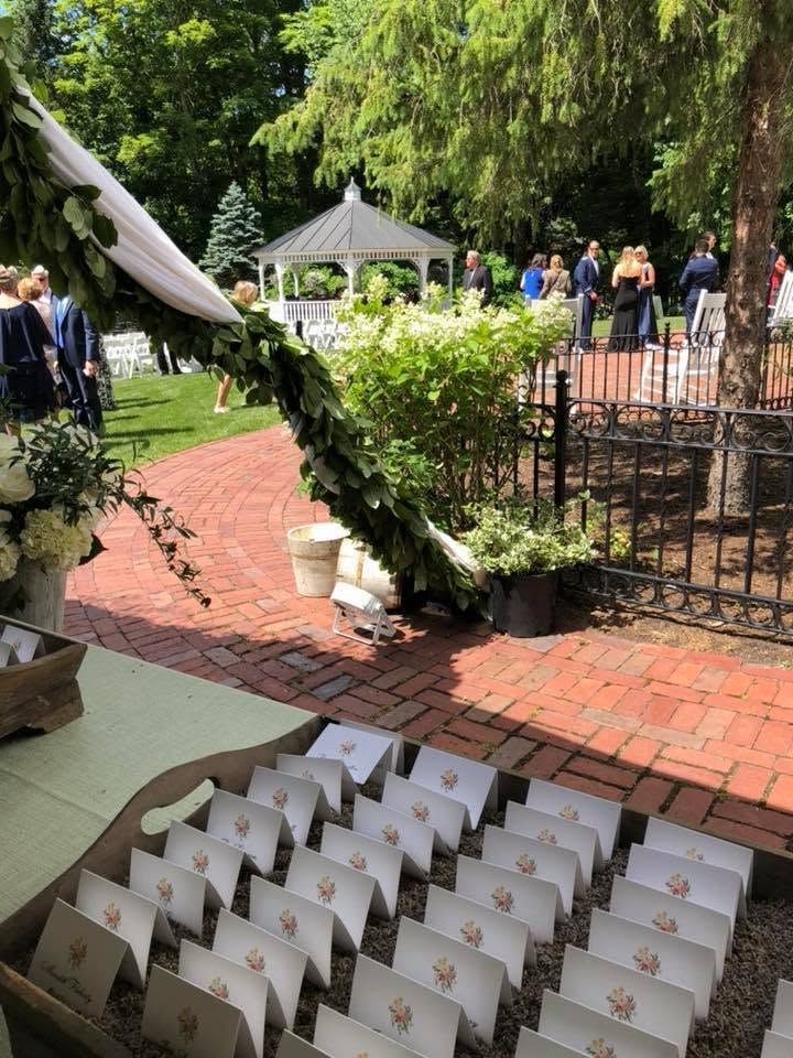 Outdoor wedding setup with white chairs, floral arch, brick path, and gazebo in a green garden