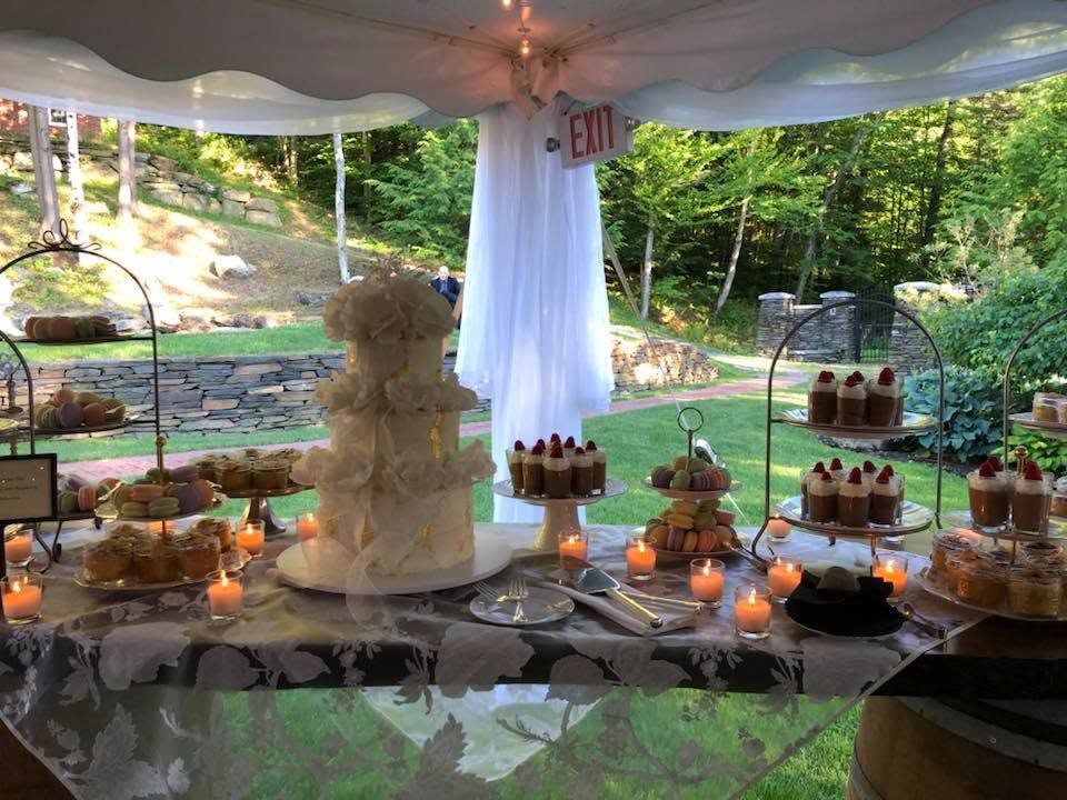 Dessert table under a tent with tiered cake, cupcakes, candles, and greenery in the background
