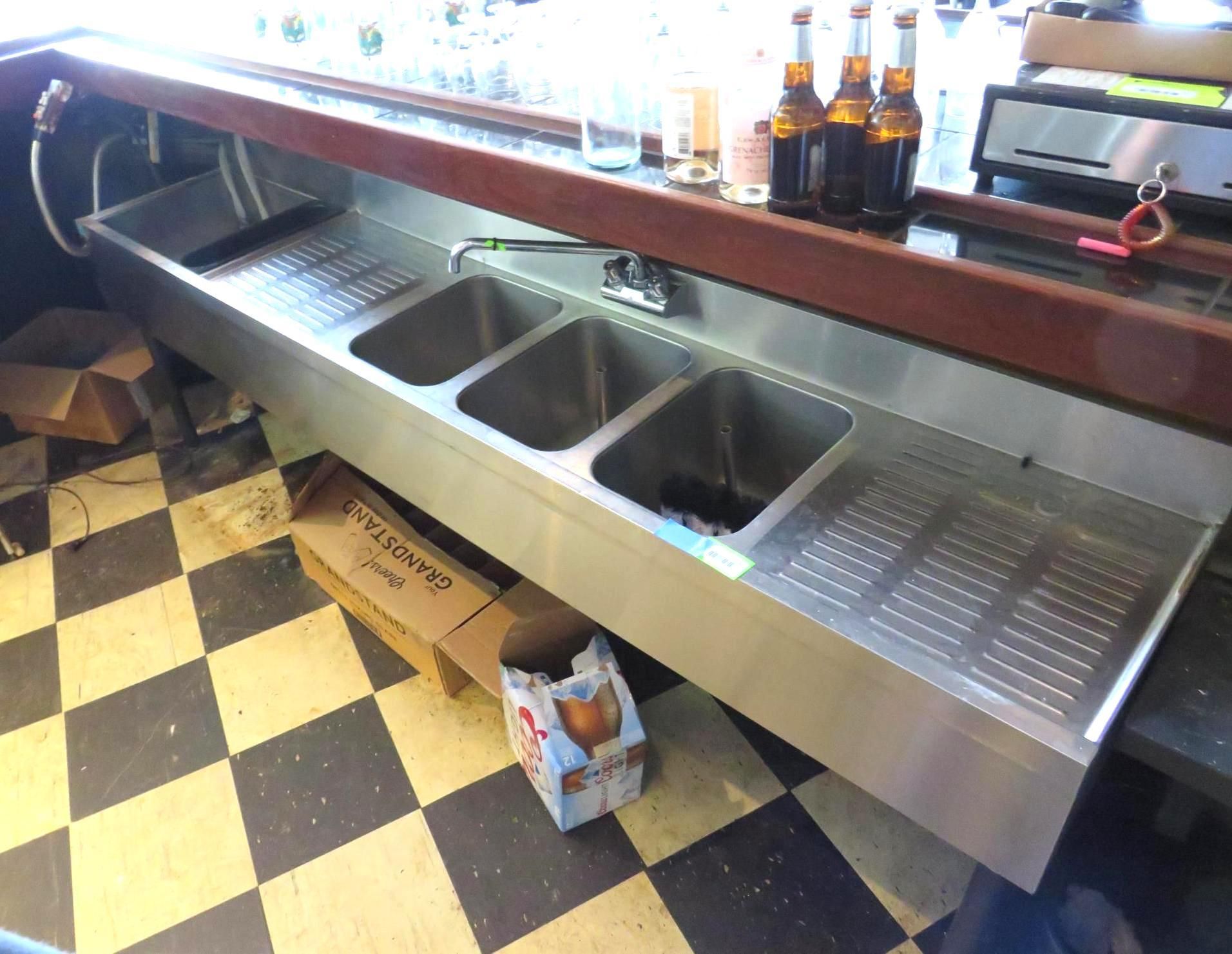 Stainless steel bar sink with three basins, behind a bar with beer bottles and a checkerboard floor.