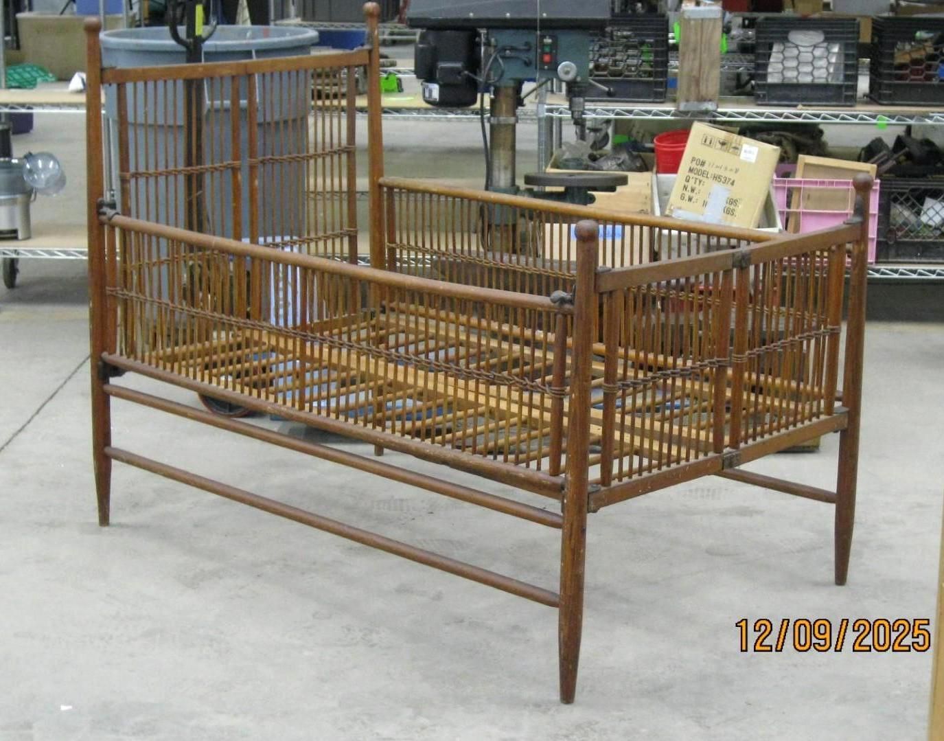 Wooden antique crib with vertical bars, brown in color, sitting on concrete floor.