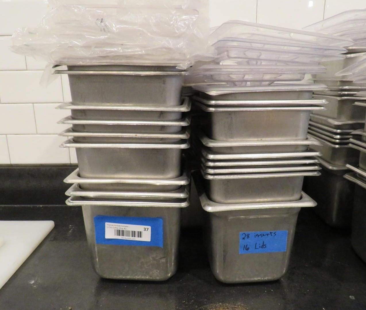 Stacks of stainless steel food containers, labeled, with clear lids. In a kitchen setting.