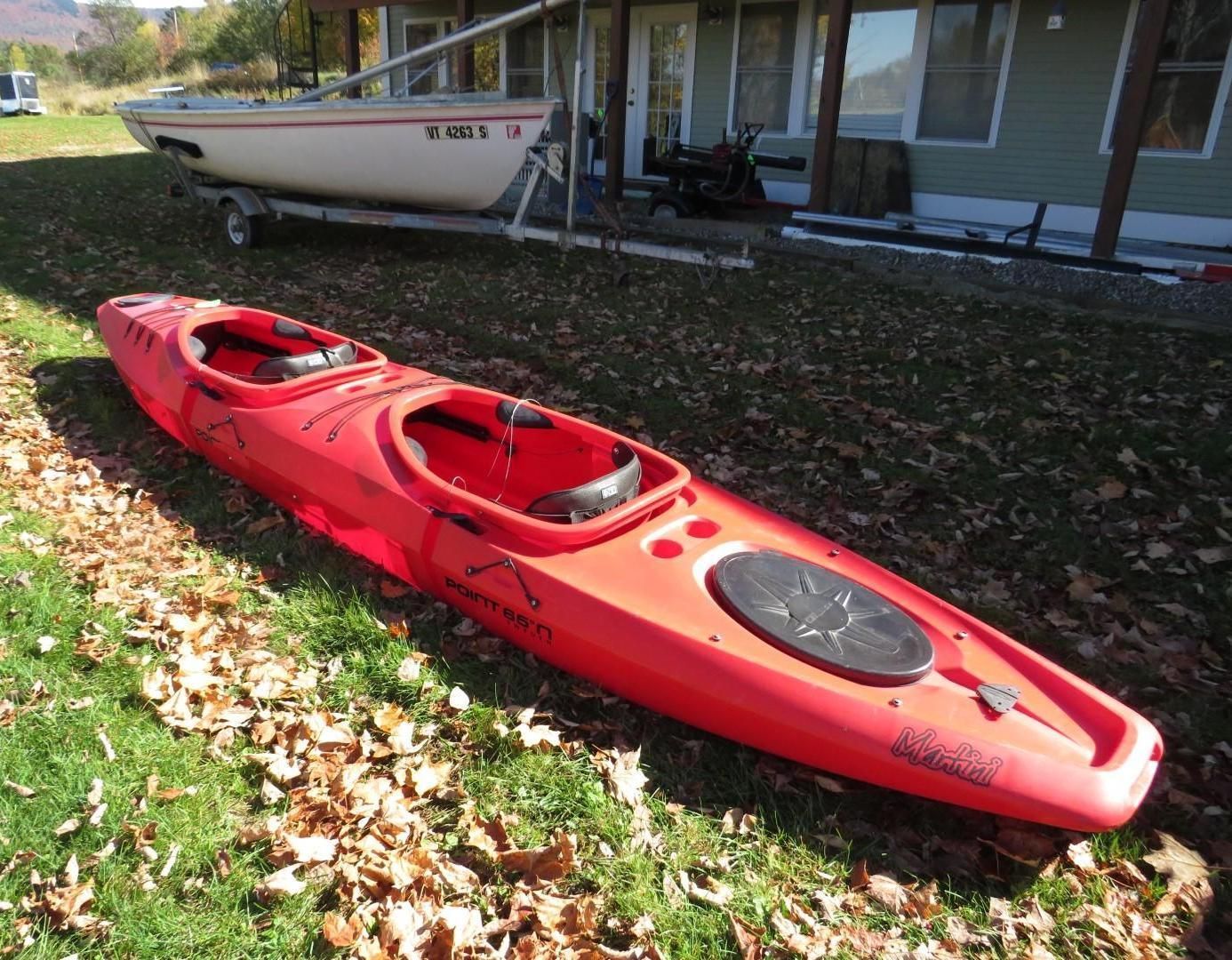 Red two-person kayak on grass, boat in background near a building.