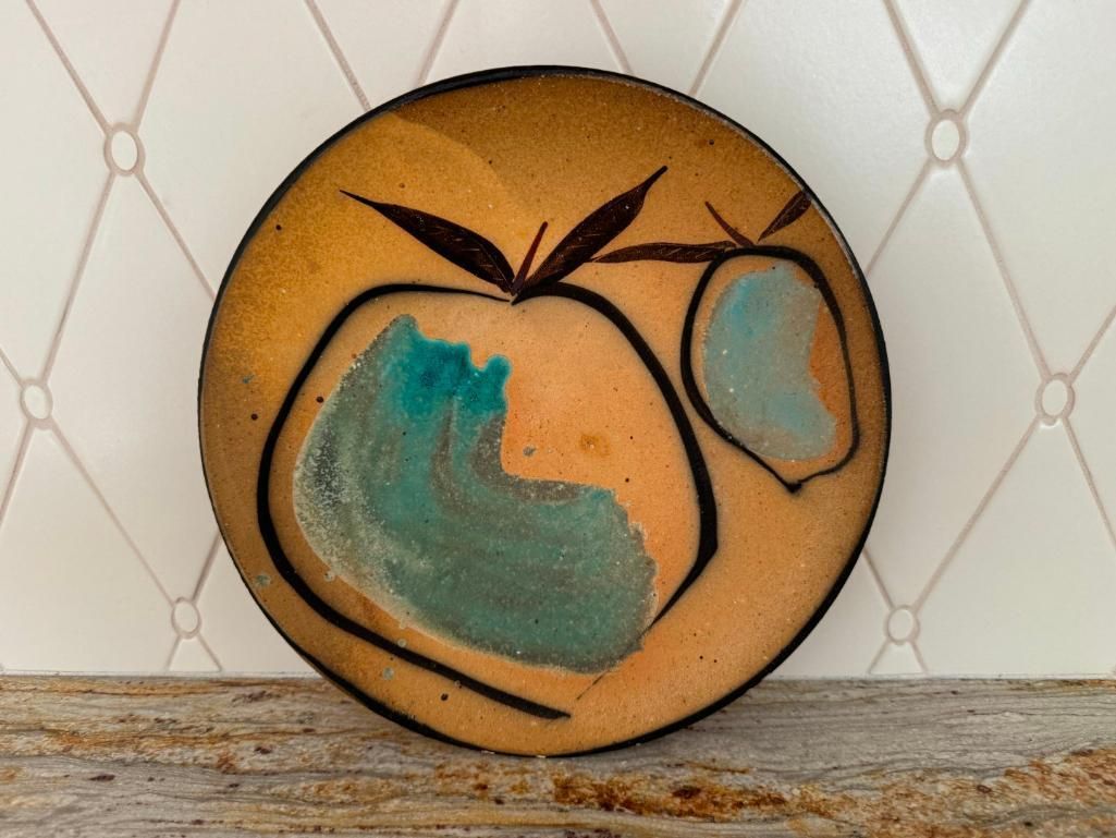Round plate with abstract fruit design in orange, brown, and turquoise against a white tile background.