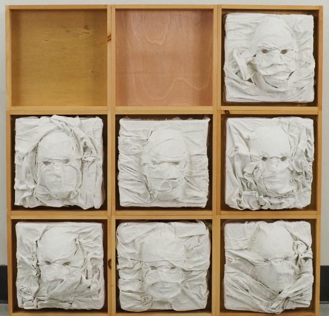 Nine-box wooden grid with 7 white sculpted faces wrapped in cloth, 2 empty boxes.