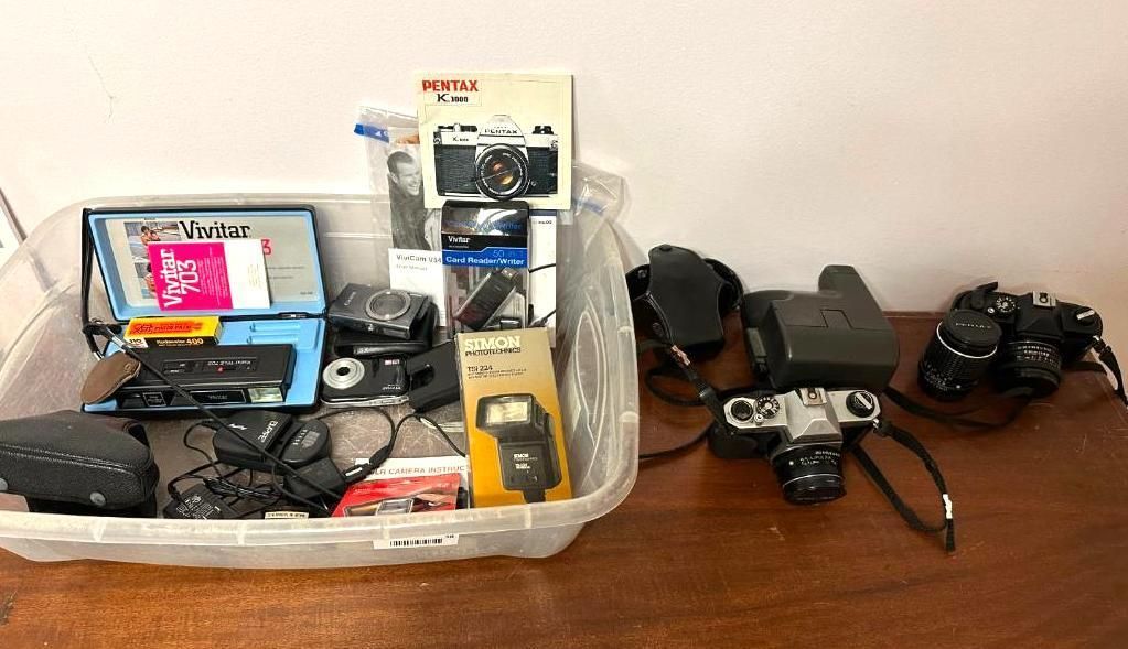 A plastic bin filled with various cameras, and other photography equipment on a wooden table.