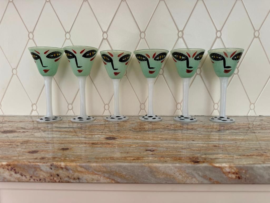 Six green face-shaped cocktail glasses with black and red details, lined up on a granite shelf.