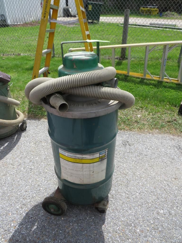 Green industrial vacuum with hose coiled on top, sitting outside on pavement.