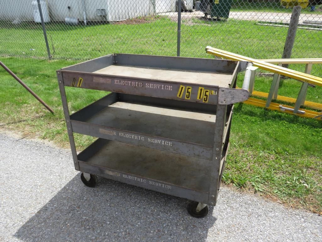 Gray metal cart with three shelves and wheels on a paved surface.