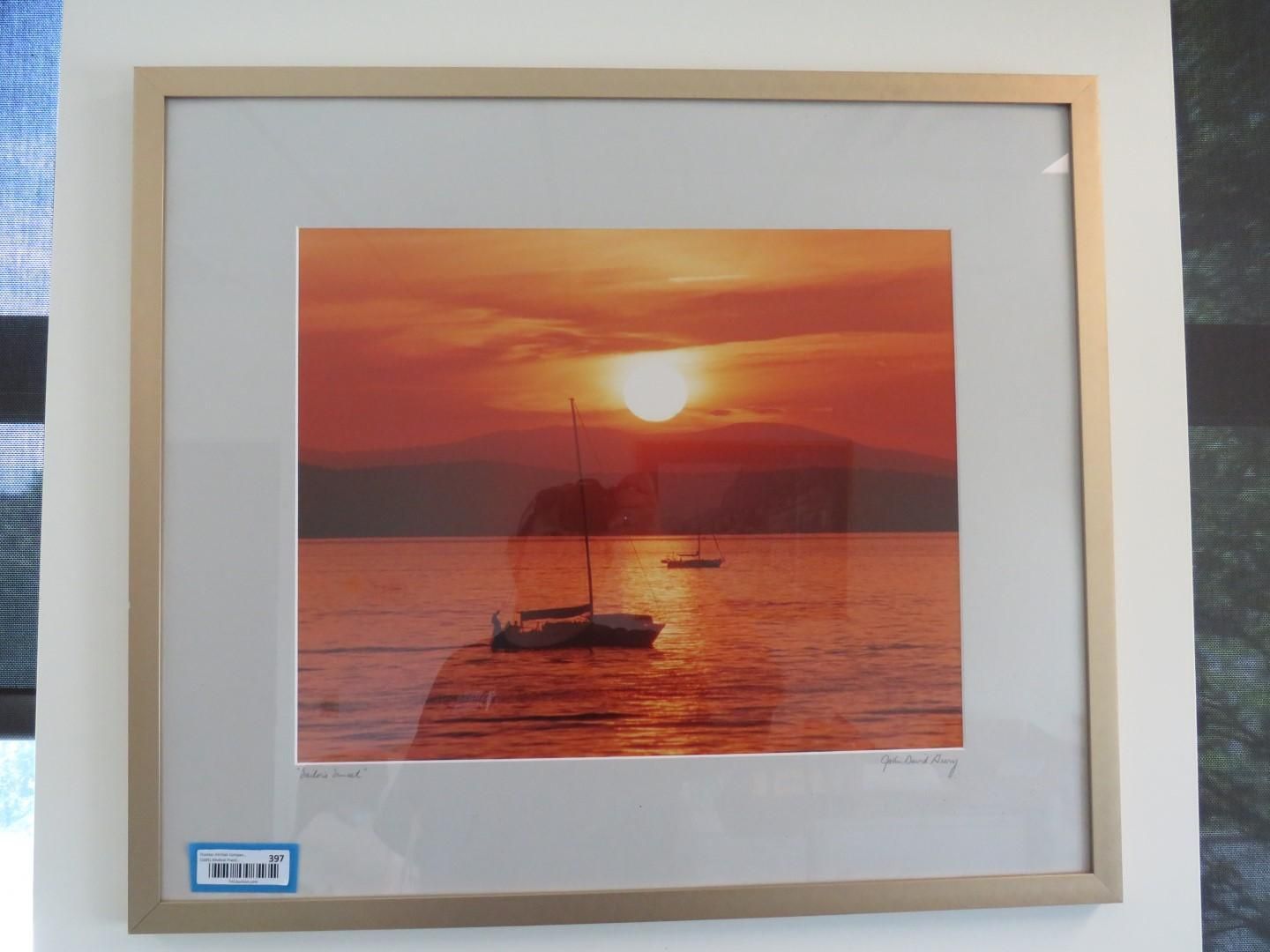 Sailboat silhouetted against a fiery sunset over calm water. Mountains in the background. Framed print.