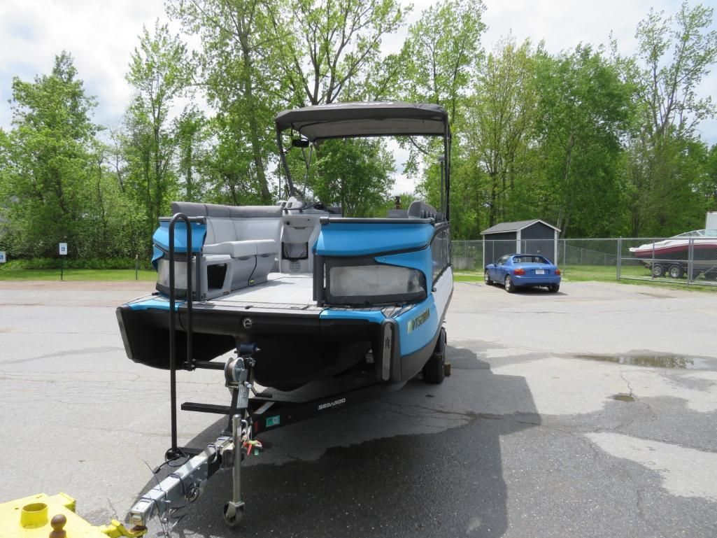 Blue and black pontoon boat on a trailer, under a canopy, parked outside on a sunny day.