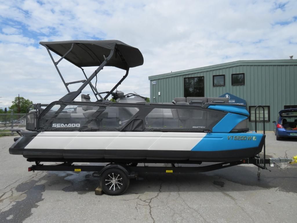 Blue and white pontoon boat on a trailer, under a black canopy. Outdoors, sunny.