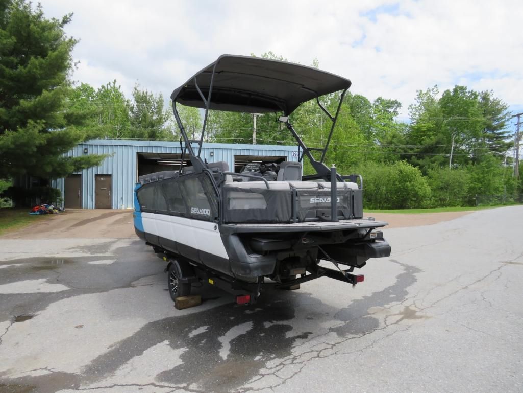 Pontoon boat on trailer, parked on a paved surface. Black canopy, blue and white hull, gray building in background.