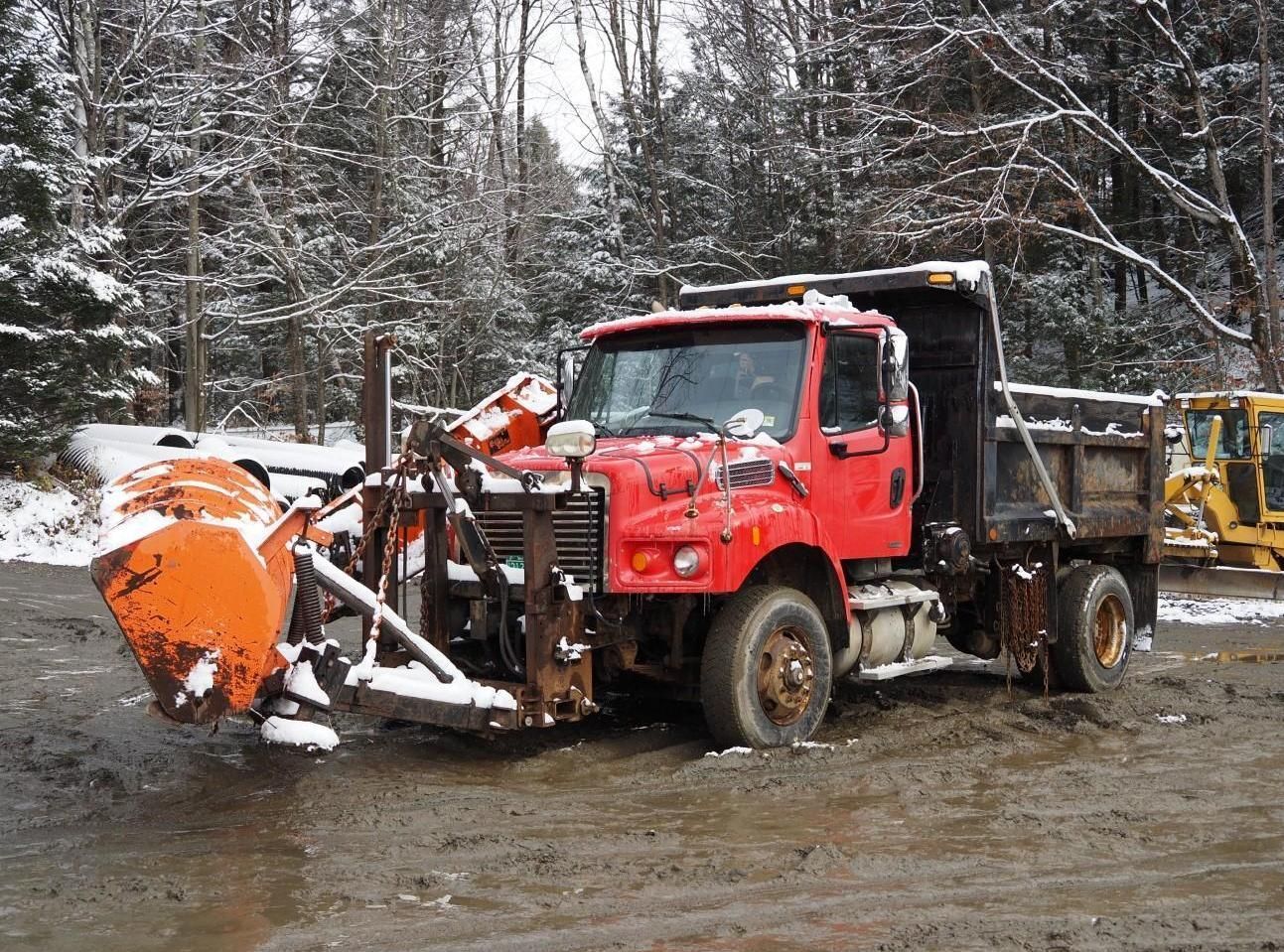 Red snowplow truck parked in muddy area, surrounded by snowy trees.