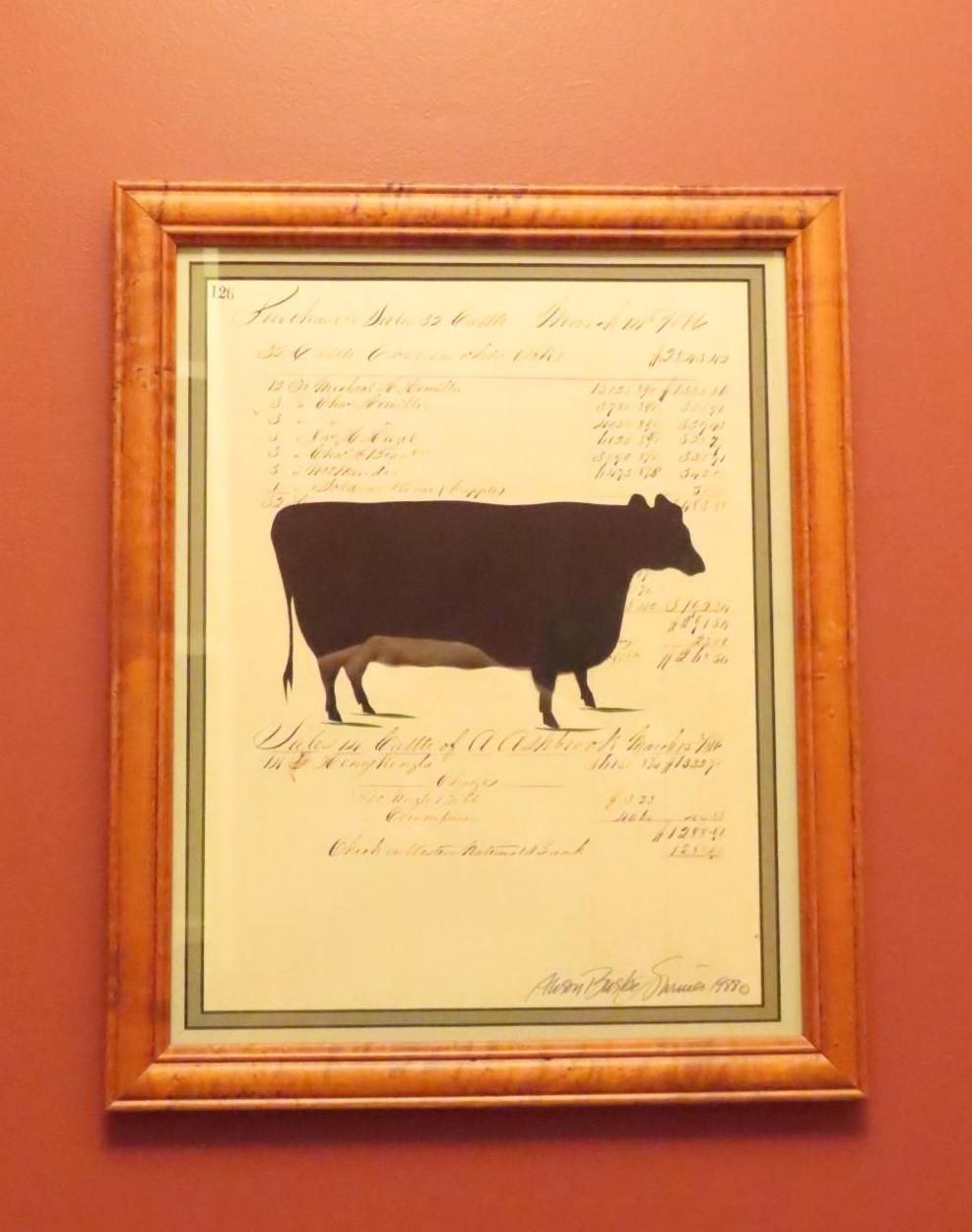 Framed artwork of a dark brown cow silhouette with handwritten notes on a cream background, against a red wall.