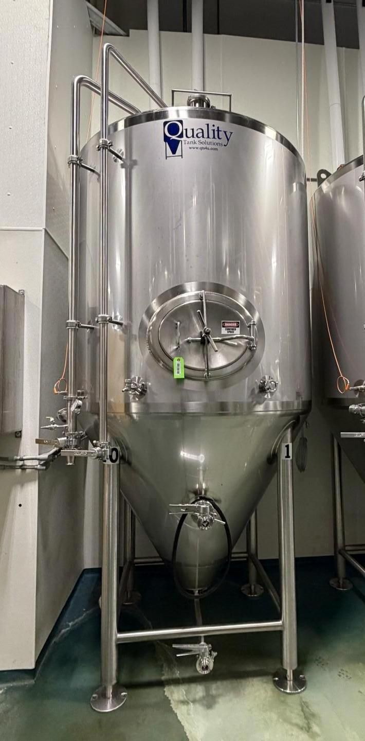 Stainless steel fermentation tank in a brewery. Cylindrical body, cone-shaped bottom. Pipes, gauges, and a large door.