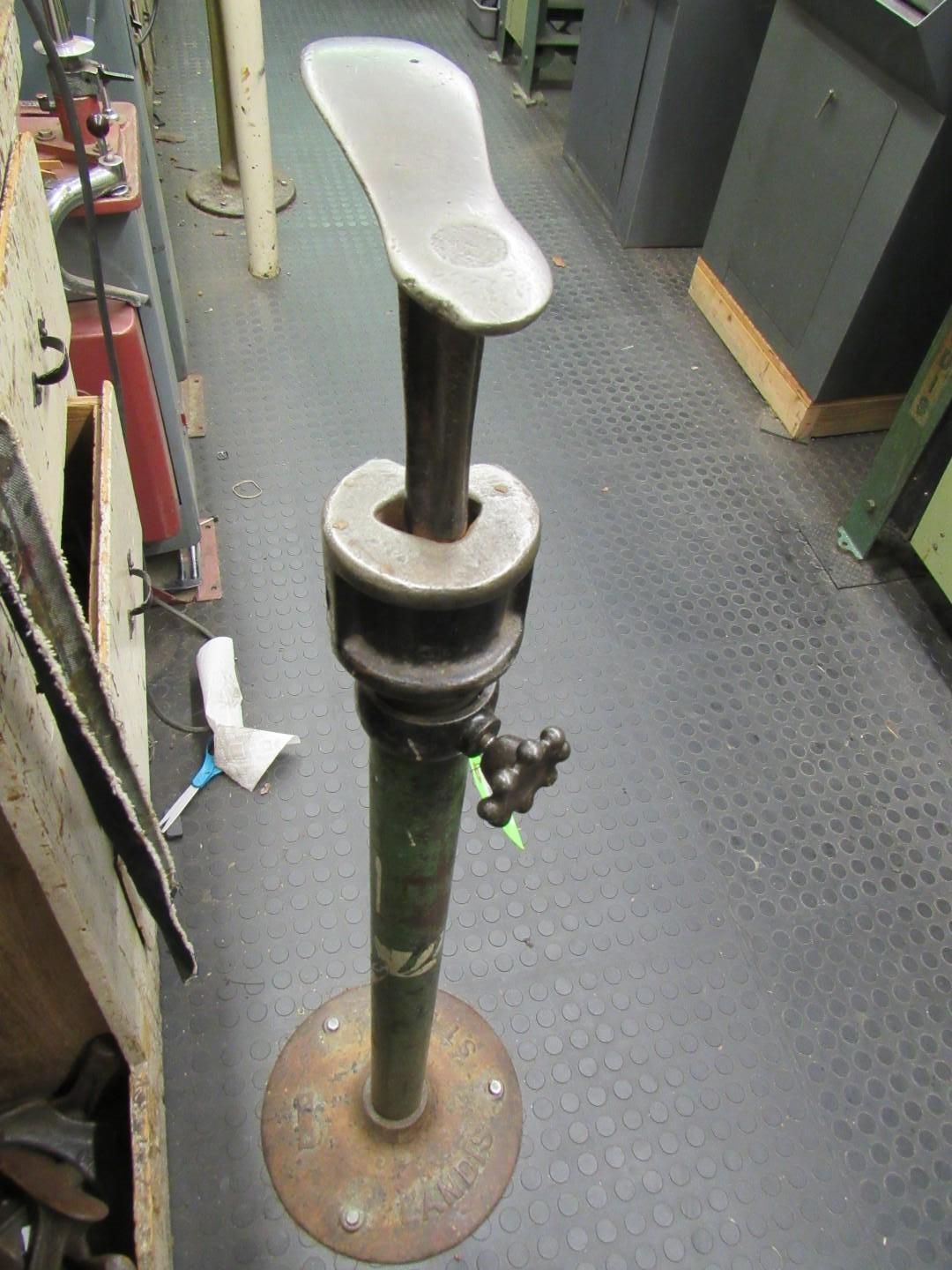 Cobbler's shoe last stand, metal, with a foot-shaped form on top, green pole, set on a workshop floor.