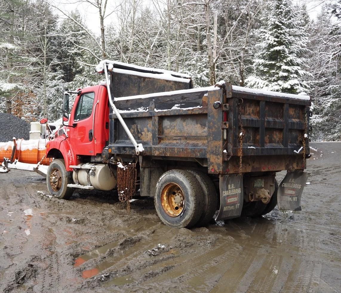 Red dump truck on a muddy road, snow-covered trees in the background.