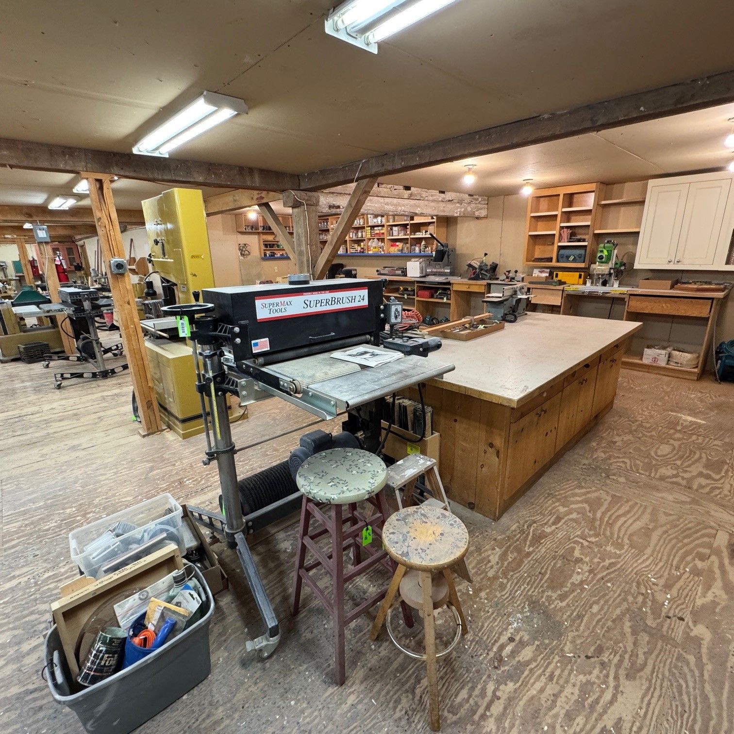 Woodworking shop with a table saw, workbenches, tools, and shelves under bright fluorescent lights