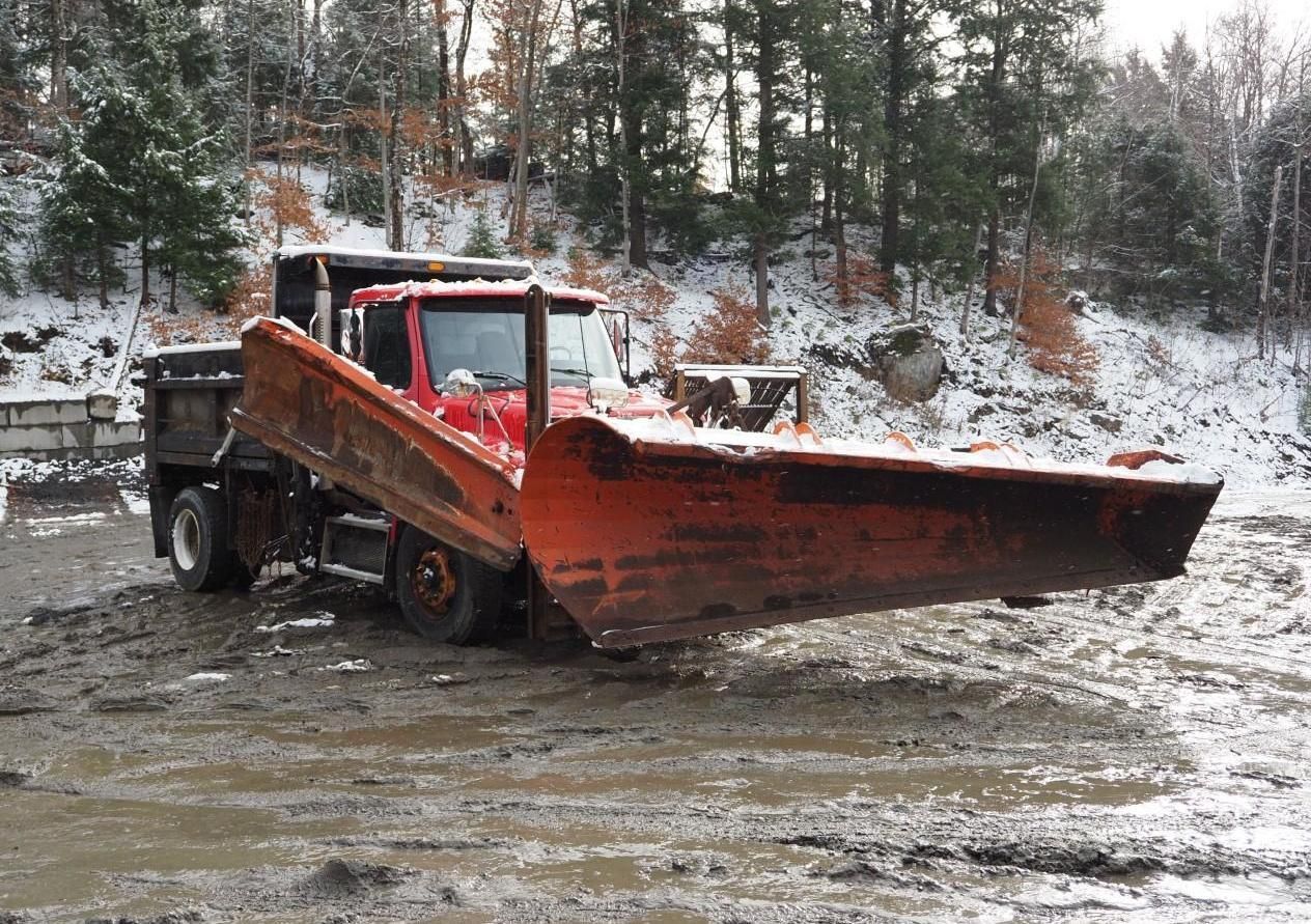 Red snowplow on muddy ground in a snowy, wooded setting.