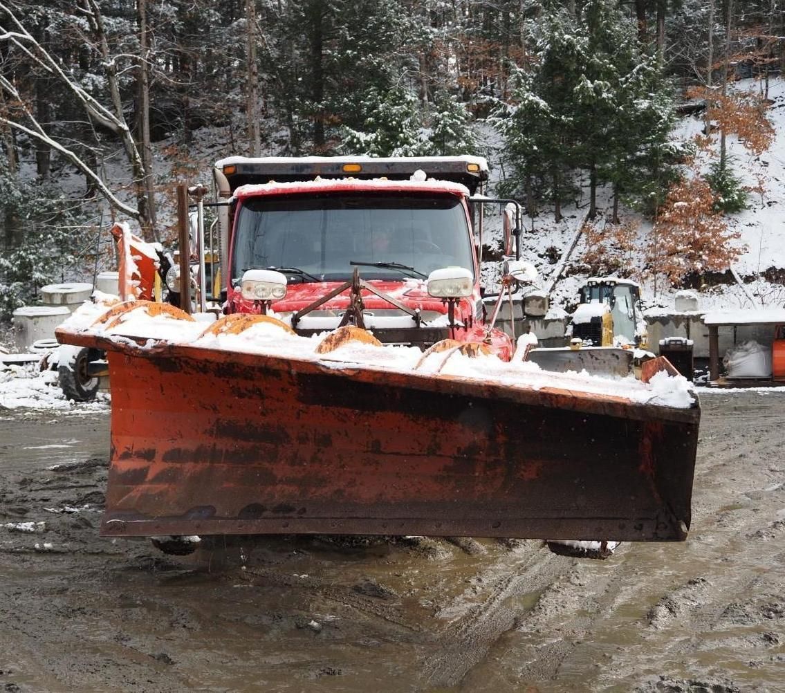 Red snowplow with raised blade clearing snow from a muddy road.