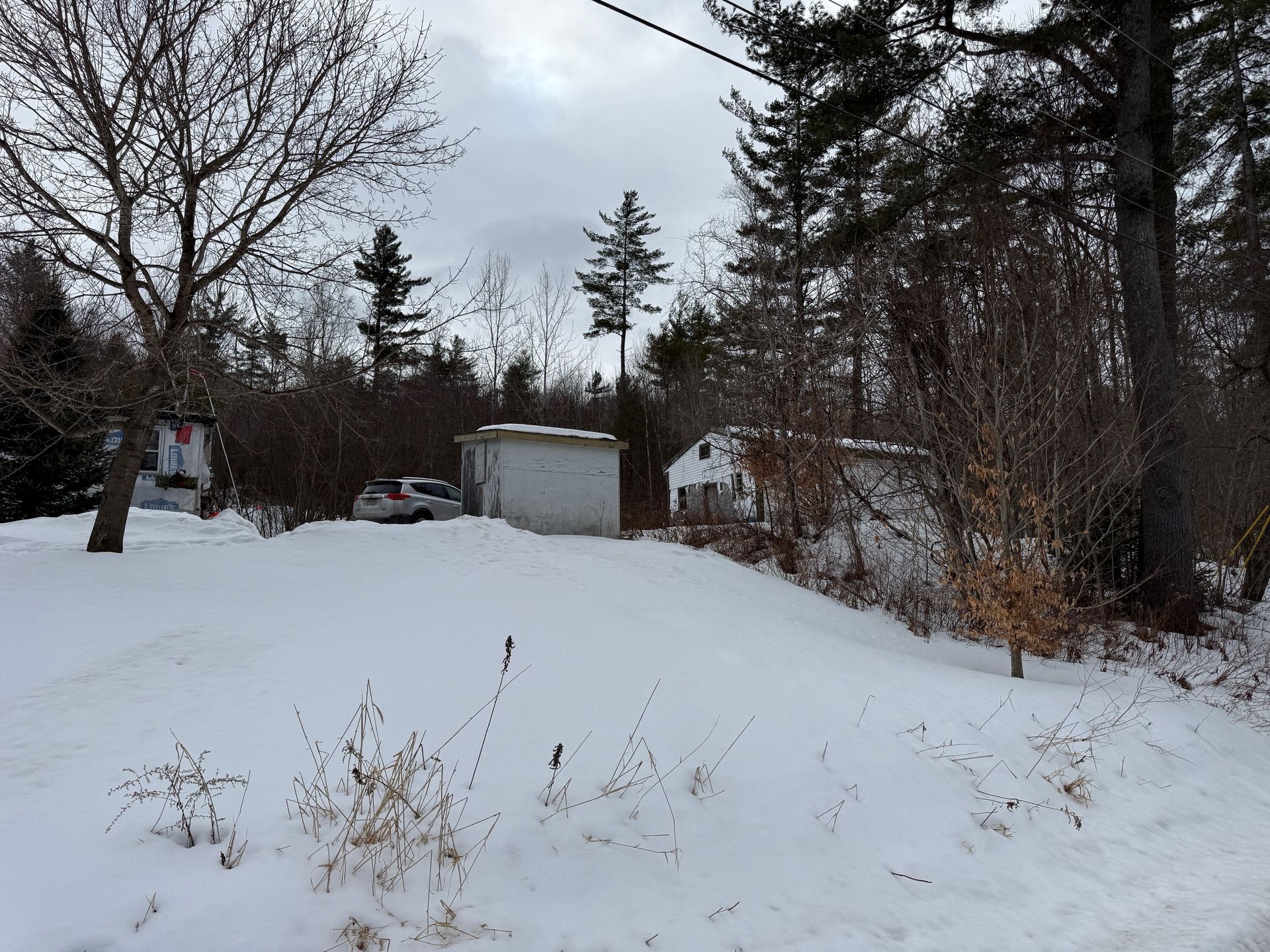 Snow-covered ground in front of two white buildings and a car, surrounded by bare trees in winter.