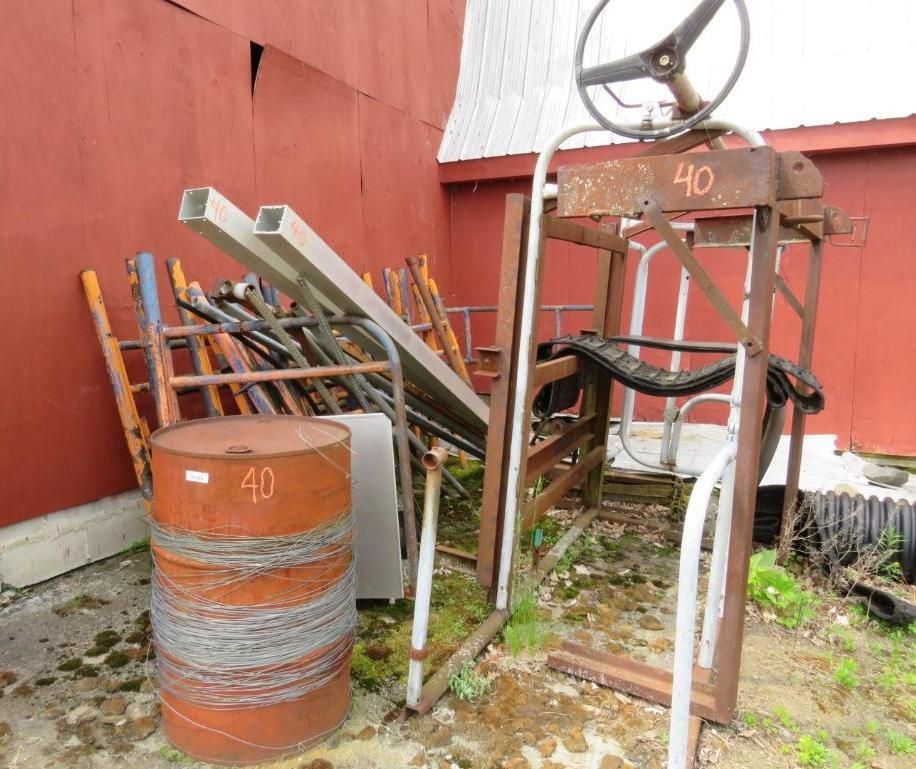 Metal equipment and barrels near a red barn. Items include a steering wheel, scrap metal, and barbed wire.