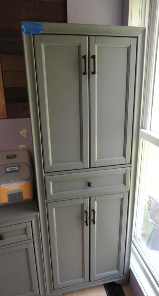 Tall, gray cabinet with two sets of doors, a drawer, and black handles, beside a window.