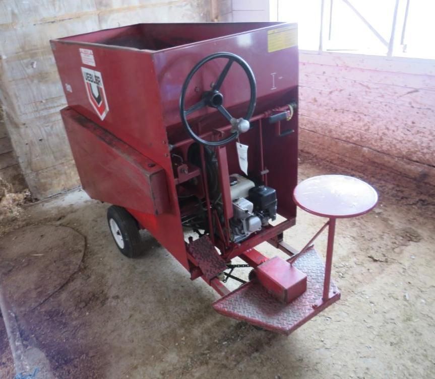 Red agricultural feed cart with steering wheel, seat, and hopper in a barn.