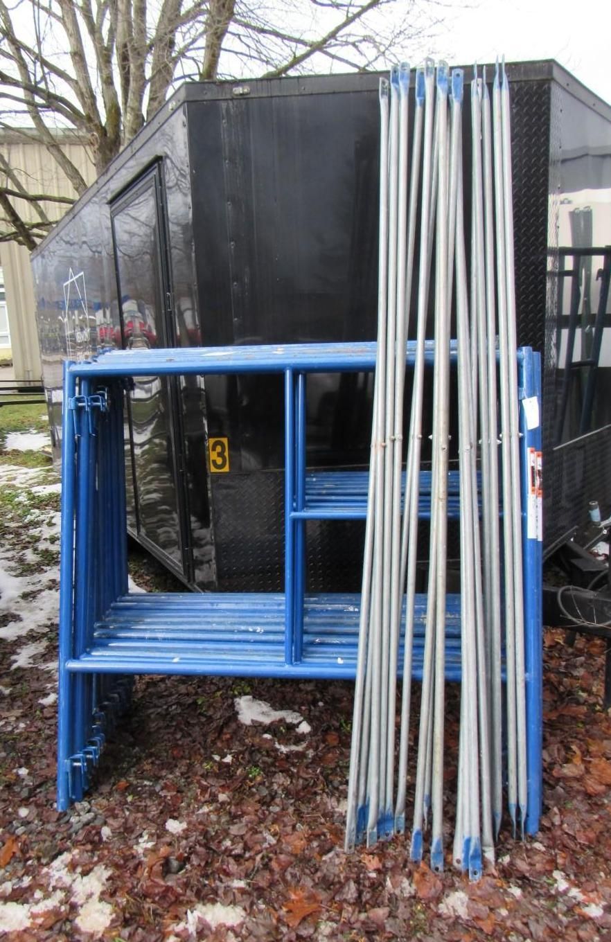 Blue scaffolding frame with stacked silver poles against a black structure outdoors.